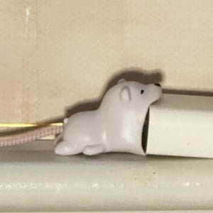May include: A white polar bear-shaped cord protector is attached to a white power adapter. The cord is pink. The items are on a white surface, with a beige wall in the background.