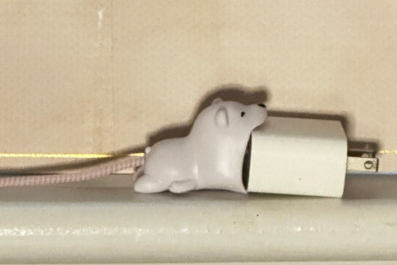 May include: A white polar bear-shaped cord protector is attached to a white power adapter. The cord is pink. The items are on a white surface, with a beige wall in the background.