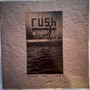 May include: A promotional poster for the band Rush, featuring the band's name in a pixelated font. The image shows a person standing on a ledge with water in the foreground. The poster is framed by a textured, gray border. The text "TOUR 1991-1992" is at the bottom.