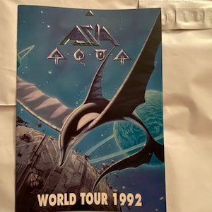 May include: A blue and white concert poster for the band Asia. The poster features a stylized image of a flying creature against a starry sky. The text "Asia" is in a large, bold font and the text "World Tour 1992" is at the bottom of the poster.