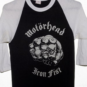 May include: Black and white baseball style t-shirt with a white graphic of a fist holding a set of pistons with skulls on them. The text "motörhead" and "Iron Fist" are printed in white on the shirt.