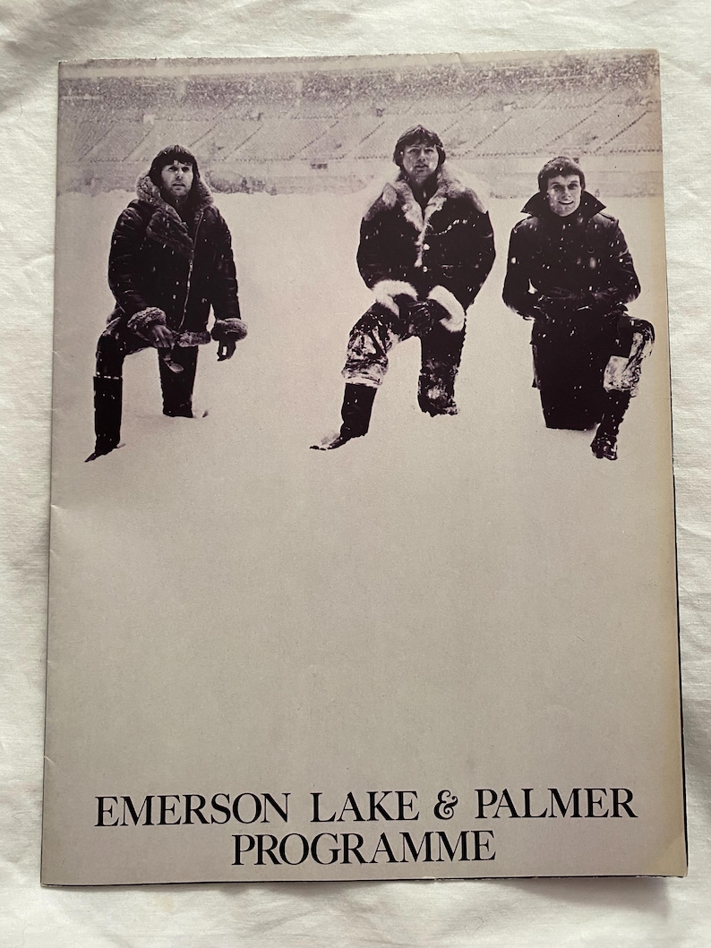 May include: A vintage concert program cover featuring three men in winter coats standing in the snow. The text "EMERSON LAKE & PALMER PROGRAMME" is printed in a serif font at the bottom of the cover.