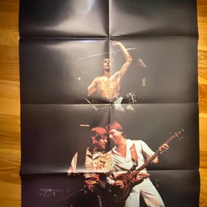 May include: A vintage concert poster for Emerson, Lake & Palmer. The poster features a shirtless man with his arm raised, and two other men playing instruments. The band's name is printed at the top in white.