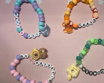 Könnte beinhalten: Fünf bunte Perlenarmbänder mit Anhängern. Die Armbänder buchstabieren die Wörter "ARF", "CRABS", "CLOUD", "MEOWWOOW" und "QT PATOO TIE".