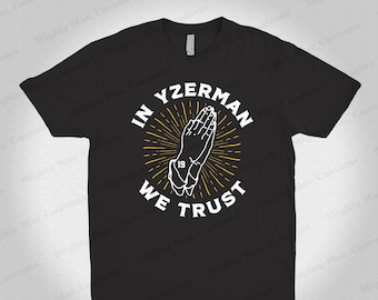 Op de afbeelding: Zwart T-shirt met een witte afbeelding van gevouwen handen met de tekst "In Yzerman We Trust" en het nummer "19" in een cirkel.
