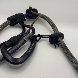 May include: A close-up of a black carabiner attached to a gray and black rope. The rope has a pattern of gray, black, and tan. A black knot and a black rubber piece are also visible. The carabiner has the text "WH-25 1767".