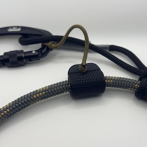 May include: A black carabiner with a locking mechanism is connected to a black and gray rope. A black textured sleeve secures the rope. A loop of olive green cord is attached.