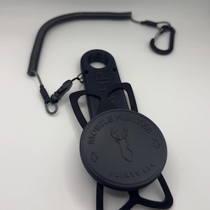 May include: A black plastic mobile phone holder featuring a circular center with the embossed text MOBILE HUNTER PRINTS LLC and a deer head silhouette. The holder includes a flexible coiled tether with a small carabiner clip attached to the top.