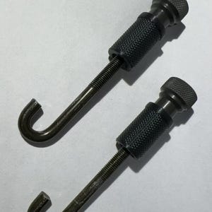 May include: Two black metal hooks with threaded shafts and knurled knobs. The hooks are J-shaped and the knobs are round.
