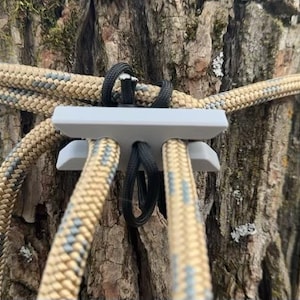 May include: A close-up of a gray plastic tree strap with four ropes threaded through it. The ropes are tan and black with a camouflage pattern.