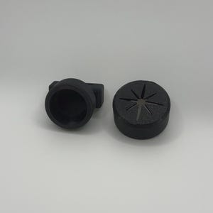 May include: Two black, 3D-printed objects. One is a cylindrical shape with a starburst pattern on top. The other is a cylindrical shape with a side extension. Both objects are on a white background.