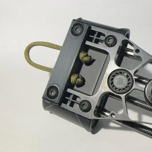 May include: A black and silver archery rest with a green cord attached. The rest has a circular design with a black and silver finish. The rest is designed to hold an arrow in place while shooting.