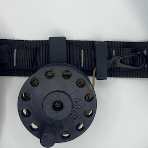 May include: A black reel with a series of holes and the letters "MHP" printed on the side. The reel is attached to a black strap with a clip and a small loop. The reel is designed to hold a cord or rope.