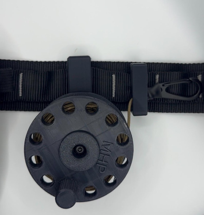 May include: A black reel with a series of holes and the letters "MHP" printed on the side. The reel is attached to a black strap with a clip and a small loop. The reel is designed to hold a cord or rope.