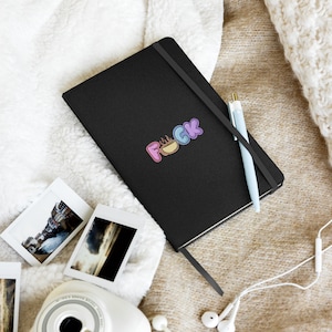 May include: Black notebook with a colorful sticker that says "F**K" with a coffee cup in the middle of the word. The notebook is on a white fuzzy surface with a pen and a pair of headphones.