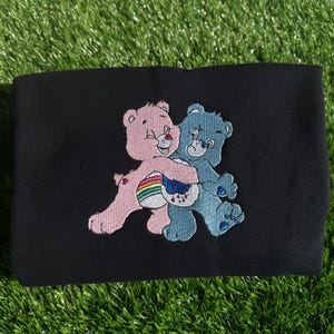 Peut inclure: Sweat-shirt noir avec un motif brodé de deux Care Bears qui s'embrassent. Le Care Bear à gauche est rose avec un arc-en-ciel et le Care Bear à droite est bleu.