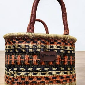 Può includere: Borsa a cestino intrecciata con un motivo geometrico arancione, nero e beige. La borsa ha manici in pelle marrone e una piccola etichetta in pelle con la scritta "Bolga Splendor". La borsa è progettata per trasportare oggetti.