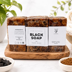 May include: Three rectangular bars of 9oz handmade black soap wrapped in white paper labels. The labels feature text including ingredients like cocoa pod, coconut oil, and shea butter, along with the words 100% natural, black soap, and organic, non-toxic, vegan.