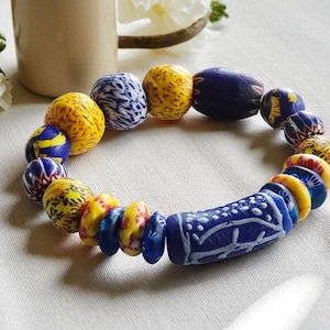 Puede incluir: Una pulsera de cuentas con una variedad de cuentas coloridas y estampadas. La pulsera incluye cuentas amarillas, azules y blancas, con una gran cuenta cilíndrica azul en el centro. Las cuentas están dispuestas en un patrón circular.