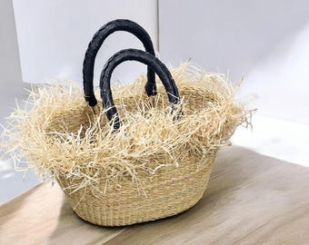 May include: Oval-shaped woven basket with black leather handles. The basket is filled with dried straw, creating a textured, natural look. The basket sits on a light-colored wooden surface.