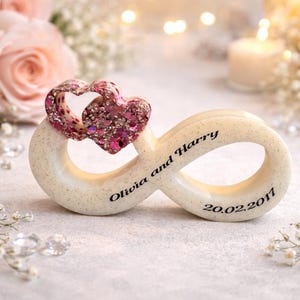 May include: A cream-coloured infinity symbol, topped with two glitter-covered pink hearts. The names "Olivia and Harry" and the date "20.02.2011" are printed on the symbol. The background features soft focus flowers and a candle.