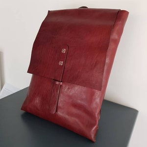 Può includere: Uno zaino in pelle bordeaux con chiusura a patta. La borsa presenta due piccoli rivetti argentati e una chiusura con cinturino in pelle. La pelle ha un aspetto strutturato. La borsa è appoggiata su una superficie blu scuro.