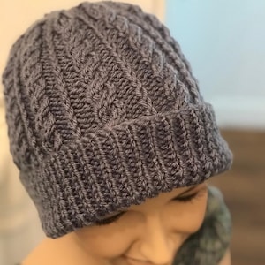Op de afbeelding: Een grijze gebreide beanie met een kabelbreipatroon. De beanie wordt gedragen op het hoofd van een persoon.