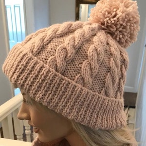 Könnte beinhalten: Eine rosa gestrickte Mütze mit einem Zopfmuster und einem flauschigen Bommel oben.