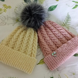 May include: Two knitted winter hats. One is pale yellow with a cable knit pattern and a matching brim. The other is pink with a cable knit pattern and a matching brim. Both hats have a large, fluffy, dark gray pom-pom on top.