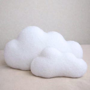 May include: Two white cloud-shaped decorative pillows. The larger pillow is positioned behind a smaller one, both made of a soft, textured material. The pillows are set against a neutral background.