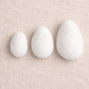 Puede incluir: Tres objetos blancos en forma de huevo de diferentes tamaños están dispuestos sobre una tela beige. Los huevos tienen un aspecto texturizado, similar al fieltro. El huevo más pequeño está a la izquierda, el más grande a la derecha.