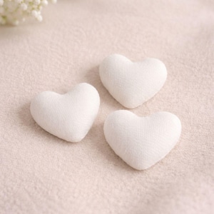 Puede incluir: Tres pequeñas decoraciones de tela en forma de corazón, de color blanco. Los corazones están dispuestos sobre una superficie beige claro, con un aspecto suave y texturizado. Los corazones están espaciados uniformemente.