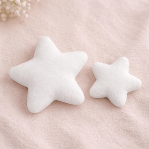 Puede incluir: Dos decoraciones de felpa blancas en forma de estrella de diferentes tamaños. Las estrellas están hechas de un material suave y texturizado y están colocadas sobre una superficie rosa claro texturizada. Las estrellas tienen un diseño simple y elegante.