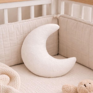 Puede incluir: Una almohada suave en forma de media luna blanca descansa en una cuna. La almohada está hecha de una tela texturizada y está rodeada por un protector de cuna y ropa de cama de color crema. Se ve un pequeño osito de peluche.