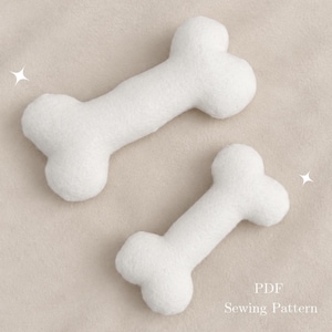 Peut inclure: Deux objets en forme d'os en peluche blanche de tailles différentes disposés en diagonale. Le texte PDF Sewing Pattern est imprimé dans le coin inférieur droit. Chaque os a une forme arrondie et segmentée avec une texture douce et duveteuse.