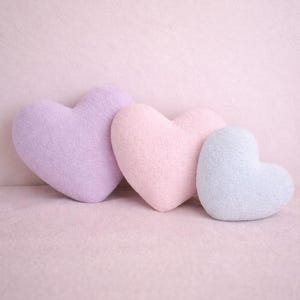 Puede incluir: Tres cojines en forma de corazón en colores pastel: lavanda, rosa y azul claro. Los cojines están dispuestos en fila, con el cojín lavanda a la izquierda y el cojín azul claro a la derecha. Están hechos de un material suave y texturizado.