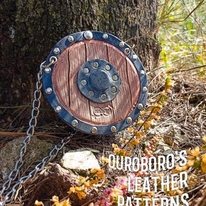 Puede incluir: Bolso redondo de cuero marrón y gris, con forma de escudo y correa de cadena. Tiene un centro con textura de madera, un borde de metal gris oscuro y un botón central de metal. El texto "OUROBORO'S LEATHER PATTERNS" es visible.
