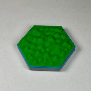 May include: A green hexagonal tile with a raised, textured surface resembling a mountain range. The tile has a blue border and a gray base.