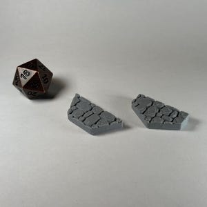 Peut inclure: Deux tuiles de terrain texturées en pierre grise pour jeux de table. Les tuiles sont en forme de coin et ont une surface rugueuse et inégale. Un dé à 20 faces brun avec des chiffres noirs est également présent sur l'image.