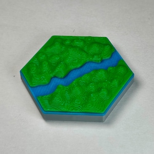 May include: A hexagonal game tile featuring a blue river flowing through green hills. The tile is made of plastic and has a textured surface.