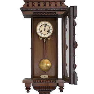 Puede incluir: Reloj de pared antiguo de madera con una esfera blanca y marcadores de hora romanos negros. El reloj tiene un péndulo y una puerta de vidrio abierta. El reloj está hecho de madera oscura con tallas intrincadas.