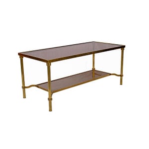 Peut inclure: Une table basse rectangulaire avec un cadre en métal couleur laiton et deux surfaces en verre. La table a une étagère inférieure et est soutenue par quatre pieds. Les surfaces en verre sont de couleur sombre et fumée.