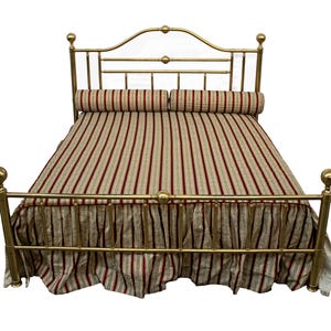 May include: A gold-colored metal bed frame with a curved headboard and footboard. The bed is made up with a striped comforter and matching bed skirt in shades of red, beige, and white. Two cylindrical pillows are placed on top.
