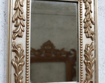 May include: A silver-toned rectangular mirror with an ornate frame featuring gold leaf accents and a beaded border. The frame has a floral design with intricate details.