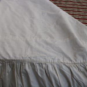 May include: Close-up of a cream-colored fabric with a pleated bottom edge. The fabric appears to be a light, possibly linen or cotton, material. The top portion is smooth, while the bottom has vertical pleats. The background shows a blue and red striped pattern.