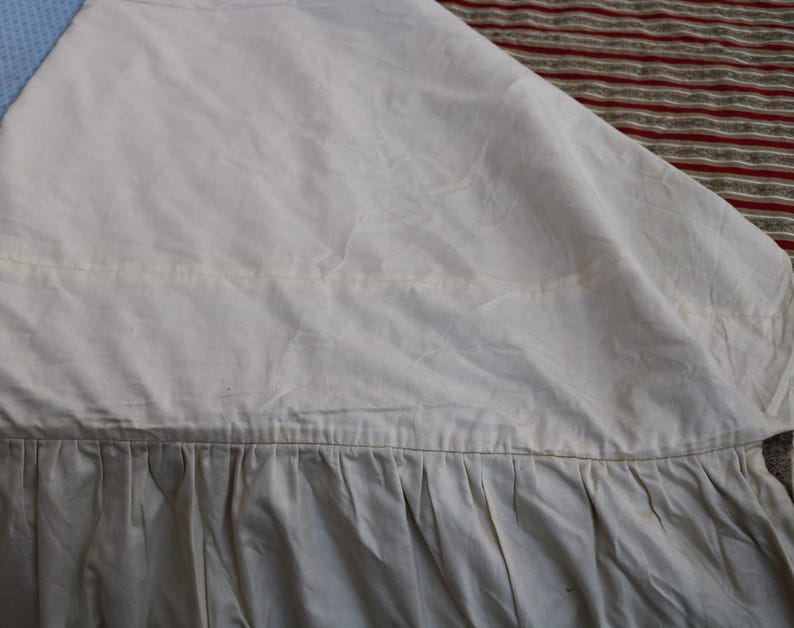 May include: Close-up of a cream-colored fabric with a pleated bottom edge. The fabric appears to be a light, possibly linen or cotton, material. The top portion is smooth, while the bottom has vertical pleats. The background shows a blue and red striped pattern.