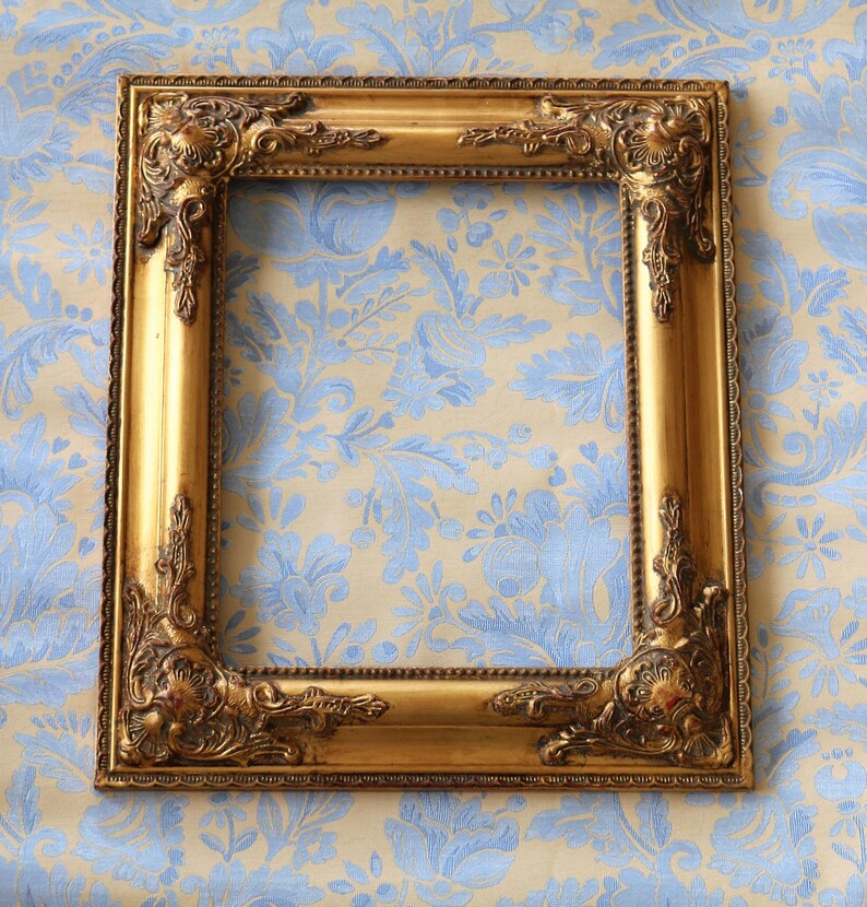 K&ouml;nnte beinhalten: Ein verzierter Bilderrahmen in Gold mit einem dekorativen Design. Der Rahmen befindet sich auf einem blau-wei&szlig; gemusterten Hintergrund.