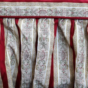 May include: Close-up of a decorative fabric with a red, cream, and gold color scheme. The fabric features vertical red stripes alternating with cream-colored stripes adorned with a gold and red floral pattern. The top edge has a decorative border.