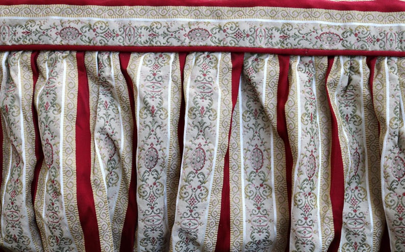 May include: Close-up of a decorative fabric with a red, cream, and gold color scheme. The fabric features vertical red stripes alternating with cream-colored stripes adorned with a gold and red floral pattern. The top edge has a decorative border.