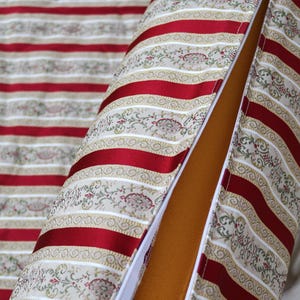 May include: A cylindrical pillow and matching fabric with a red and beige striped pattern. The pillow has a red stripe design with floral accents and a zipper. The interior is a solid tan color.